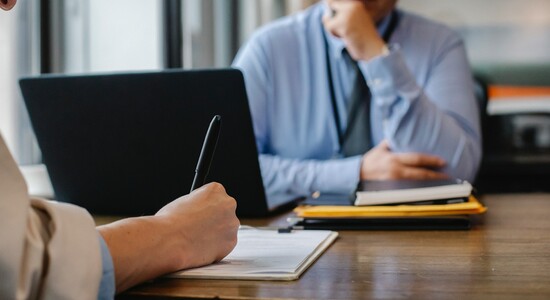 Besprechung zweier Personen wobei die eine vorm Laptop sitzt und die andere mitschreibt